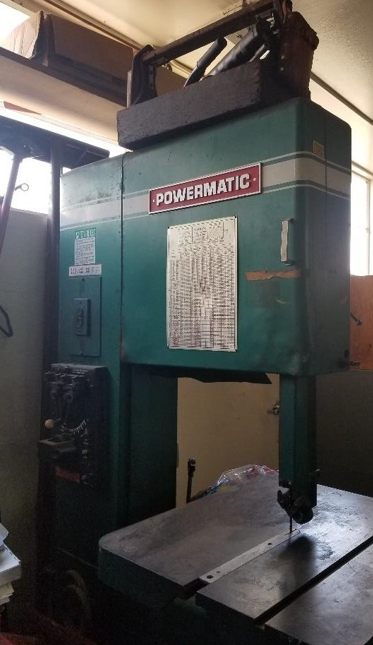 Photograph of a large green Powermatic bandsaw machine used for cutting wood or metal, positioned indoors near a window. The machine features a red and white Powermatic label, a detailed chart or instruction sheet attached to the front, and a flat cutting table with a vertical blade.